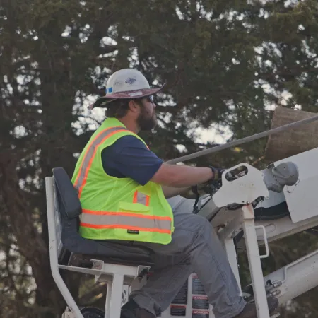 Construction worker operating a crane or lift machine outdoors wearing safety vest and helmet near trees.