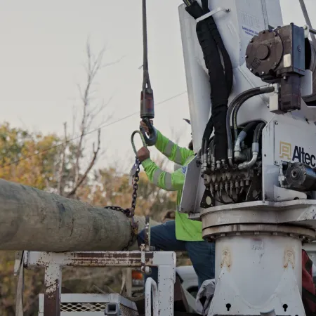 Utility workers in safety gear operating equipment to lift and install a large wooden utility pole outdoors.