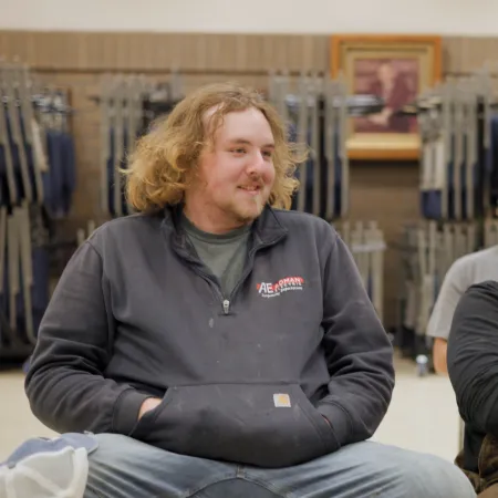 Two men sitting indoors smiling and chatting in a casual group setting with folded chairs in the background