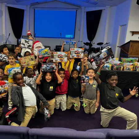 Group of excited children holding toy boxes and smiling indoors, with a person dressed as Santa in the background.