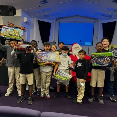 Group of children holding LEGO sets, posing with Santa Claus in a room with a projector screen and dark curtains.