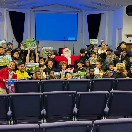 Group of children with Santa Claus holding toys and gifts in a festive indoor setting with chairs and projection screen.