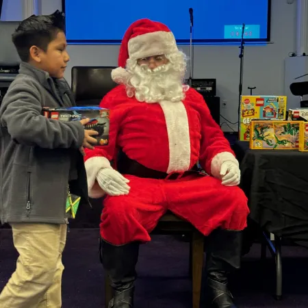 Child holding a LEGO toy box walking past a seated Santa Claus in a festive indoor setting with gifts on a table.