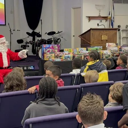 Santa Claus engaging with children seated in rows during a holiday event with toys and decorations on stage.