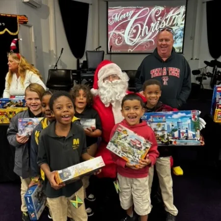 Children holding Christmas gifts posed with Santa Claus and a man in a festive indoor setting with Merry Christmas sign.