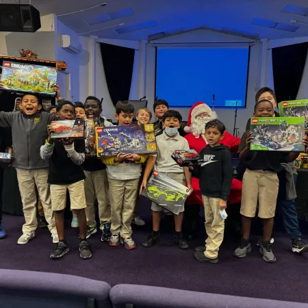 Group of children holding LEGO sets and gifts with Santa Claus in a festive event inside a hall.
