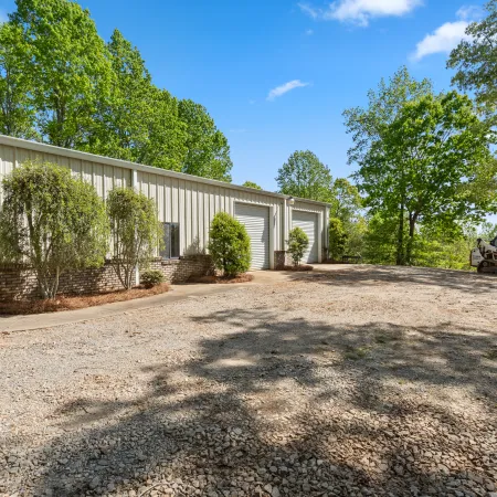 Metal workshop building with brick foundation surrounded by trees and gravel driveway on a sunny day