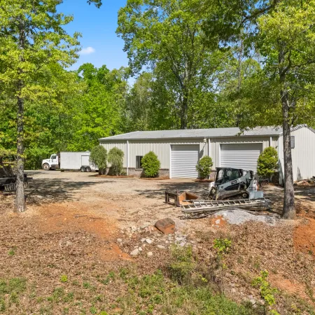 Metal storage building with three large garage doors surrounded by trees and construction equipment on dirt ground