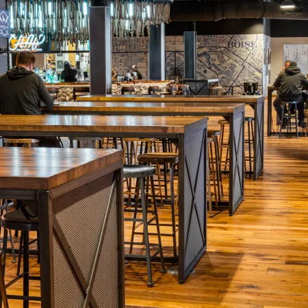 Interior of a modern bar with wooden high tables, stools, and patrons sitting near the bar area under hanging lights.