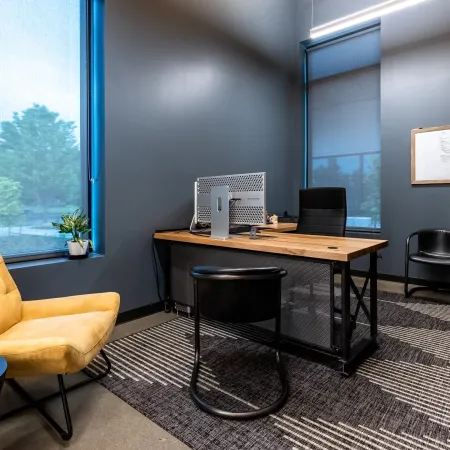 Modern office with a wooden desk, computer, black and yellow chairs, large windows, and indoor plants.