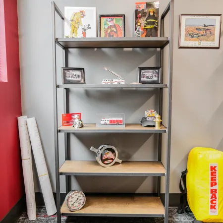Black metal shelf displaying firefighter memorabilia including photos, toy fire trucks, helmet, and 'Keep Back' safety equipment.