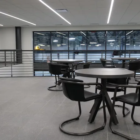 Modern indoor seating area with round tables and black chairs overlooking a large industrial space through glass windows.