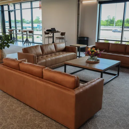 Modern office lounge with three brown leather sofas, wooden table, and large windows overlooking highway.