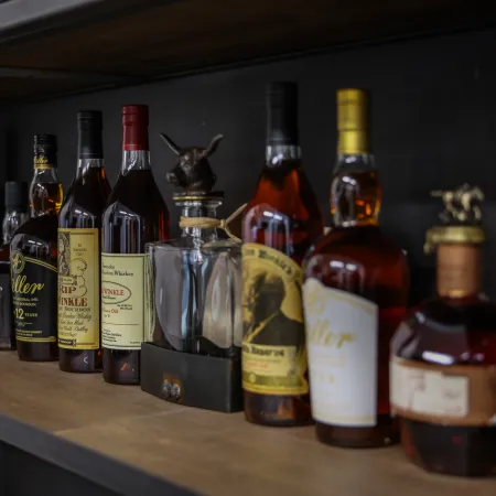 Row of assorted whiskey and bourbon bottles displayed on a wooden shelf in a bar setting.