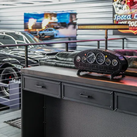 Modern garage interior with black car, vintage speedometer clock, leather couch, and neon Speed Demon sign.