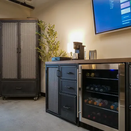 Modern kitchenette with metal cabinets, a glass-door beverage fridge, coffee maker, and potted plant under a wall-mounted screen.