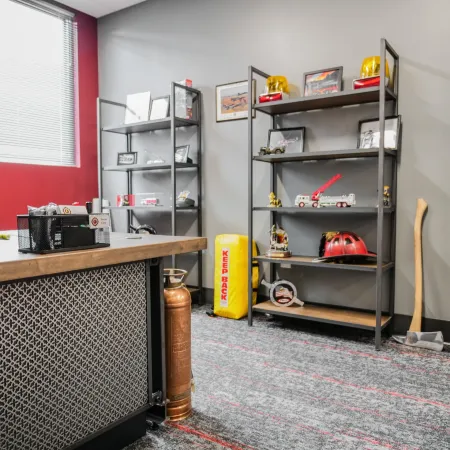 Office corner with fire safety equipment, decorative shelves, fire helmets, and safety tools on carpeted floor.