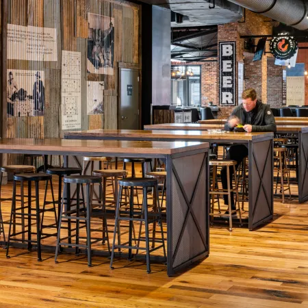 Modern industrial-style bar interior with wooden floors, metal stools, and a man sitting alone eating.