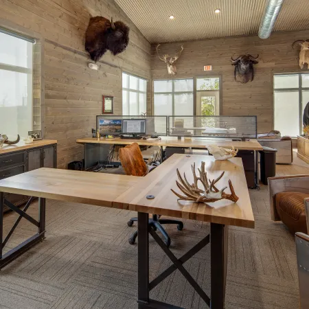 Rustic office with wooden desks, leather chairs, animal mounts on walls, and natural light through large windows.