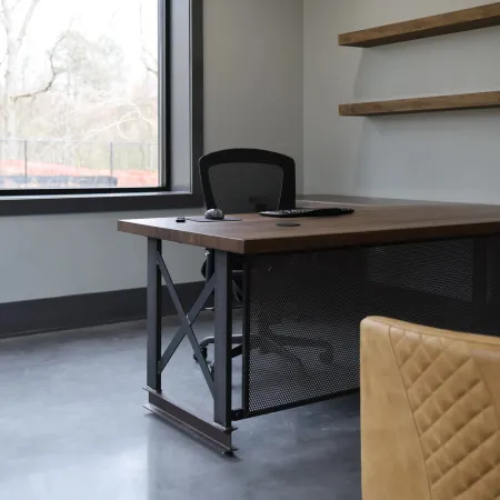 Modern office space with wooden desk, mesh office chair, leather chair, large window, and floating shelves.