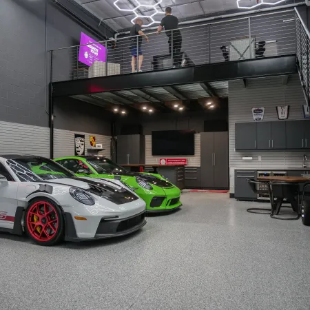 Modern garage with two sports cars, gray floor, black cabinets, and a mezzanine with two people talking above.