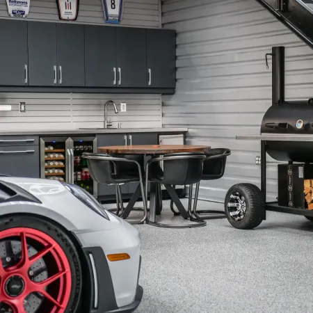 Modern garage corner with sleek cabinets, round table with chairs, smoker grill, and a sports car front wheel.