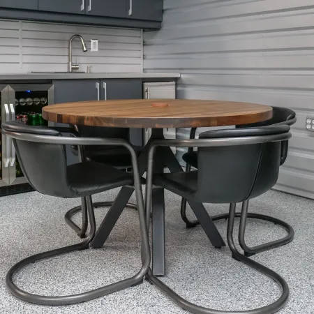 Round wooden dining table with four black leather chairs in a modern garage setting with beverage fridge and sink.