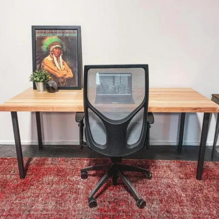 Minimalist workspace with a wooden desk, black mesh chair, framed Native American poster, plant, and red rug.