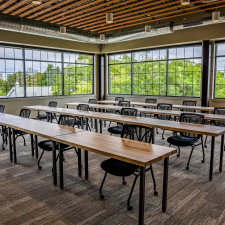 Bright modern classroom with wooden tables, black chairs, large windows, and carpeted floor in natural light.