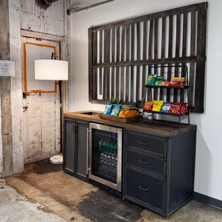 Industrial-style snack station with chips, soda fridge, wooden countertop, exposed concrete, and a floor lamp in a cozy corner.