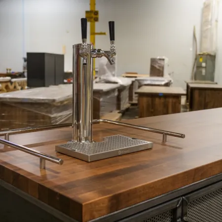 Double tap beer dispenser with stainless steel handles on a wooden bar counter in a workshop setting.