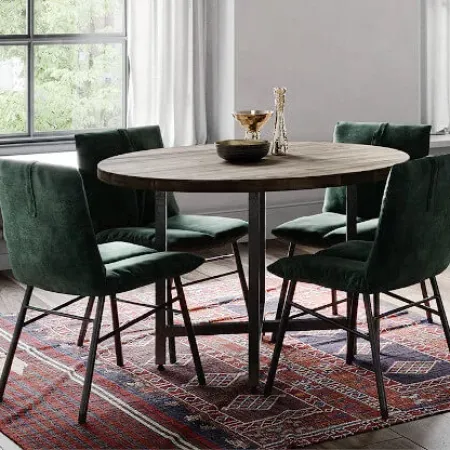 Round wooden dining table with four green velvet chairs on a patterned rug near large windows.