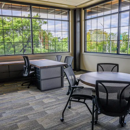 Bright modern office with large windows, wooden desks, round meeting table, and mesh office chairs.