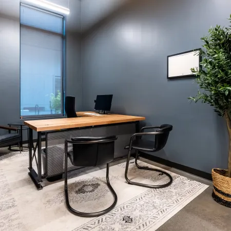 Modern office with wooden desk, black chairs, gray walls, large window, patterned rug, and potted plant in woven basket.