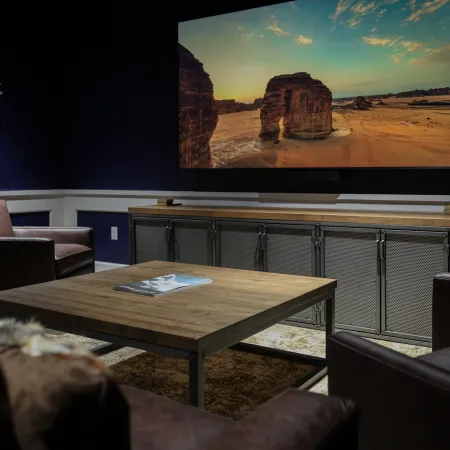 Cozy home theater room with leather chairs facing a large screen showing a desert landscape at sunset.