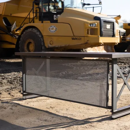 Metal construction desk with perforated panel in front of heavy yellow construction equipment on dirt ground.