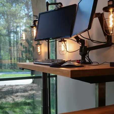 Modern standing desk setup with dual monitors, keyboard, mouse, and industrial-style wall lamps in natural light.