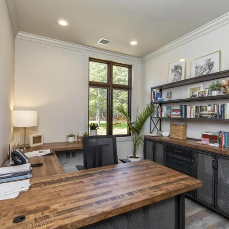 Modern home office with wooden corner desk, black mesh chair, bookshelves, and large window with greenery outside