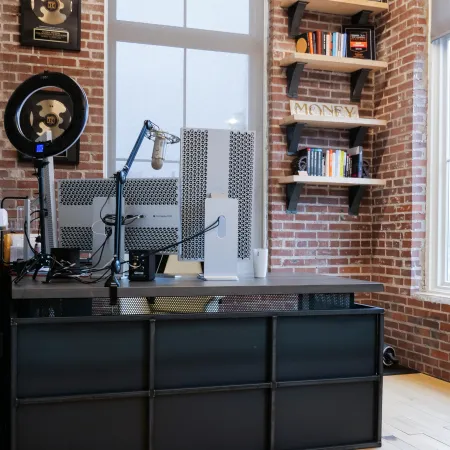 Modern office space with brick walls, dual monitors, podcast microphone, ring light, and wooden shelves with awards and books.