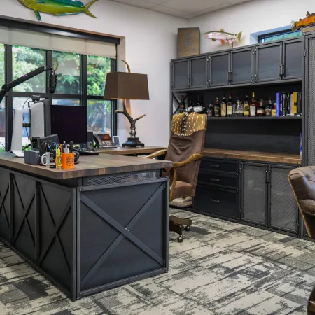 Modern office with black metal desk, leather chairs, wooden shelves, and large window with greenery outside.