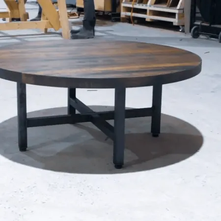 Round wooden coffee table with metal legs on a concrete floor in a workshop setting.