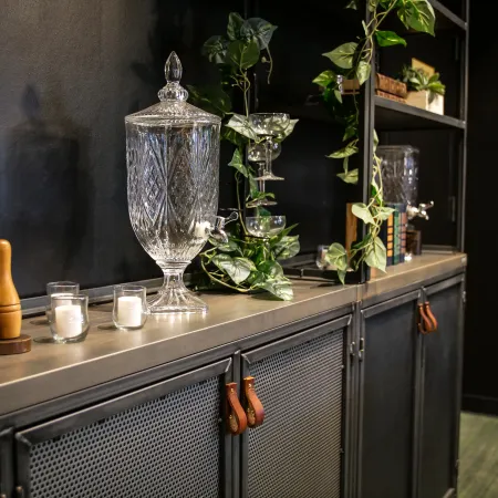 Modern black metal cabinet with leather handles decorated with crystal beverage dispenser, candles, and green plants.