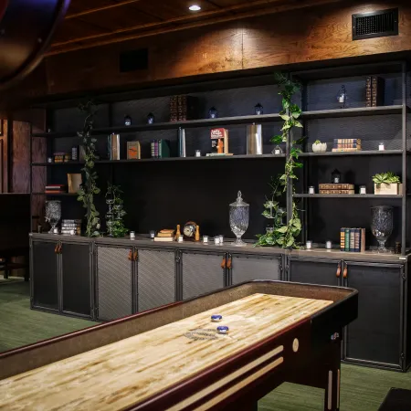 Cozy game room with shuffleboard table, wooden shelves with books, plants, and decorative glass jars under warm lighting.