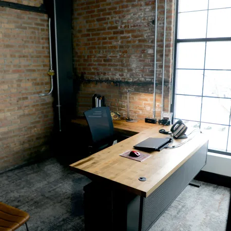 Modern office workspace with wooden desk, laptop, phone, ergonomic chair, and large window with brick walls.