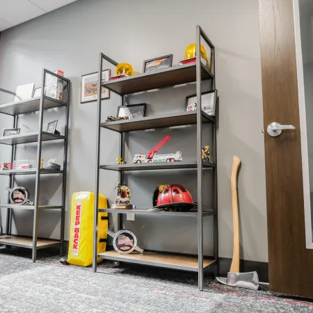 Modern room with metal shelves displaying firefighter helmets and toys, an axe and a copper container near a wooden door.
