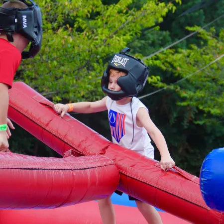 Two children wearing helmets play with large padded jousting sticks outdoors surrounded by greenery.