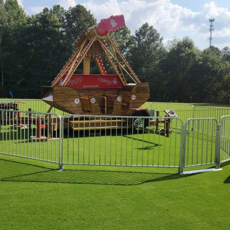 Small pirate ship ride enclosed by metal fences on green grass field under blue sky with trees in background