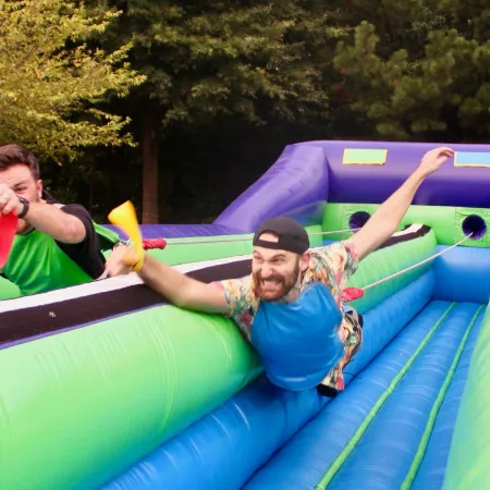 Two men compete enthusiastically on an inflatable obstacle course outdoors surrounded by trees under bright daylight.