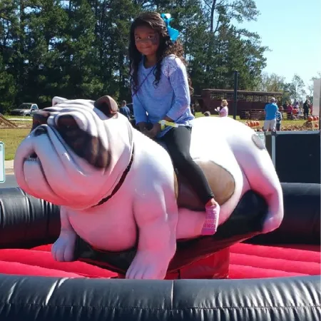 Young girl riding a mechanical bulldog at an outdoor fall festival with trees and people in the background