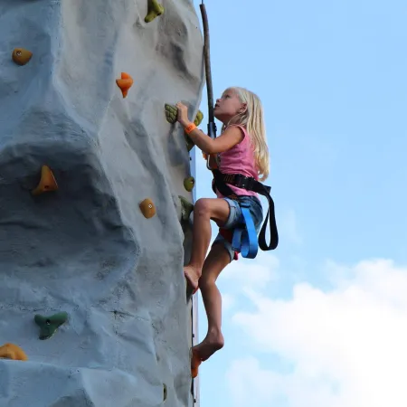 Young girl in pink shirt climbing an artificial rock wall outdoors with safety harness against blue sky.